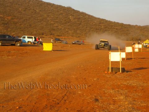 1990 Ford Bronco - off-road buggy with a roof light bar trails dust toward white course-marker signs as pickups and a yellow flag line the desert pit road.