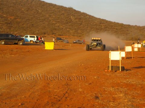 1990 Ford Bronco - off-road buggy kicks up dust along a dirt road past white course-marker signs, a yellow flag and parked pickups in the desert pit area.