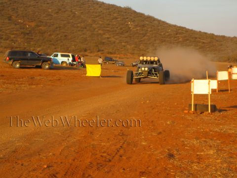 1990 Ford Bronco - off-road buggy with four roof lights trails dust past parked SUVs, a yellow course flag and white marker signs in the desert at sunset.
