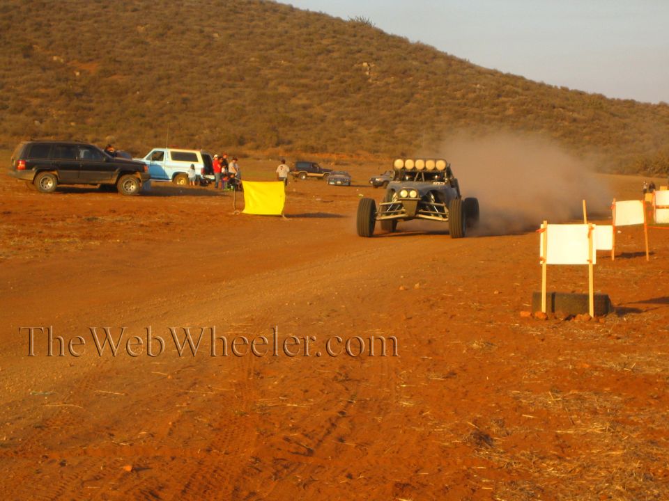 Off-road buggy with four roof lights races past parked SUVs and course signs