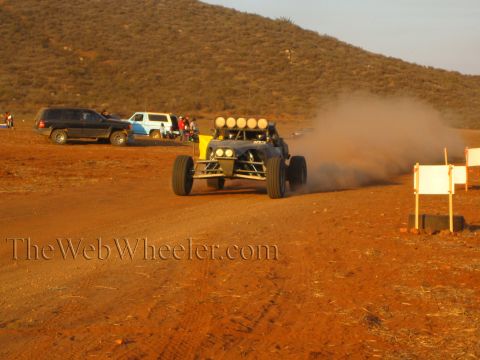 1990 Ford Bronco - off-road buggy with white roof lamps powers through a cloud of dust on a desert road past parked SUVs and course markers at sunset.