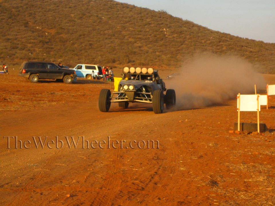 Off-road buggy with white roof lights powers through desert dust past pit trucks