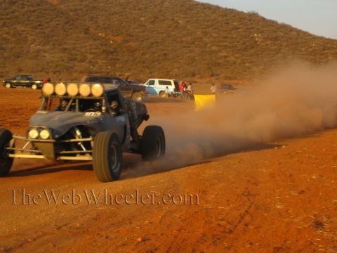 1990 Ford Bronco - dark off-road buggy with a roof light cluster slides through heavy desert dust past parked vehicles, a yellow flag and spectators.