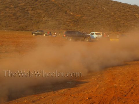 1990 Ford Bronco - Pickups and SUVs sit parked in a dusty desert pit area with several people standing nearby, partly obscured by a thick haze of blowing dust.