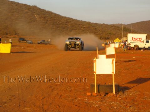 1990 Ford Bronco - off-road race truck trails dust toward white course-marker signs while a TSCO box trailer and pit canopy sit in the desert pit area at suns…