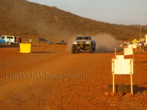 1990 Ford Bronco - off-road trophy truck trails a cloud of dust down a desert dirt course as parked vehicles and spectators line the background.