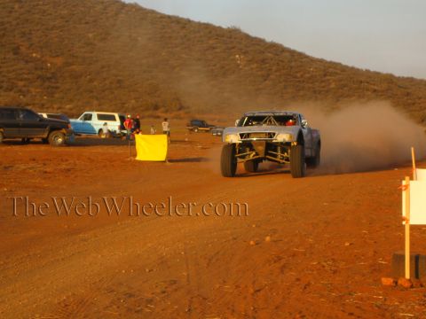 1990 Ford Bronco - silver off-road race truck races along a dusty desert track with a yellow course marker and parked trucks beside a brushy hill.