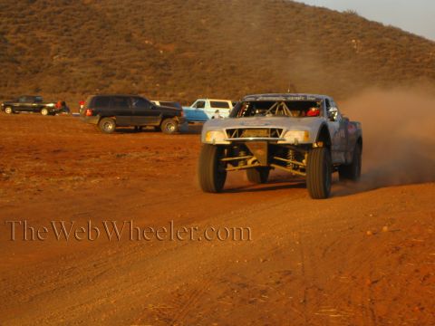 1990 Ford Bronco - off-road race truck with exposed headlights drives down a dirt track as SUVs and a pickup sit parked along the course.