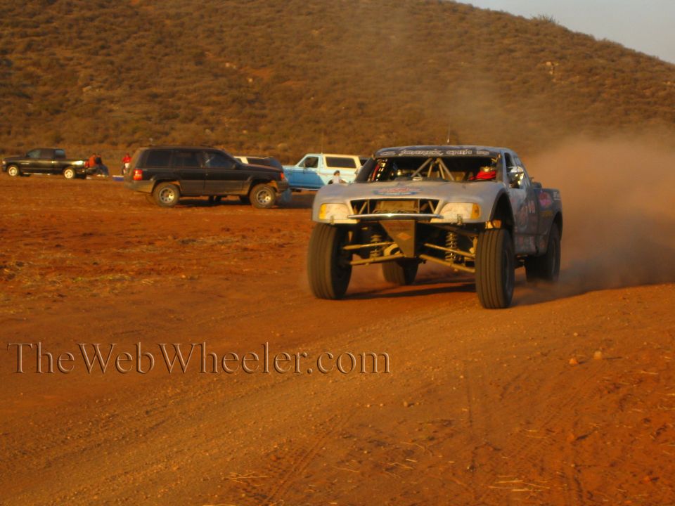 Off-road race truck with open headlights driving on desert dirt past parked SUVs