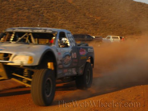 1990 Ford Bronco - sticker-plastered off-road race truck leans through a turn throwing dust, with a driver visible in the caged cockpit.