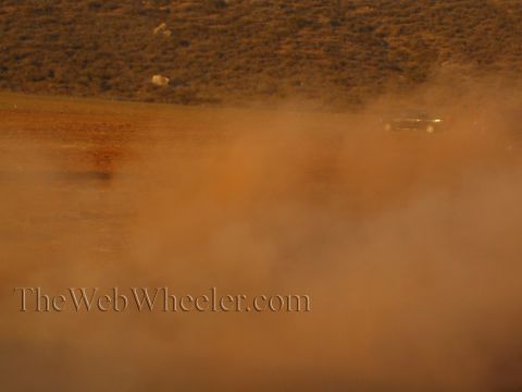 1990 Ford Bronco - vehicle is barely visible on a brushy desert hillside, nearly hidden behind a thick rising cloud of reddish dust.