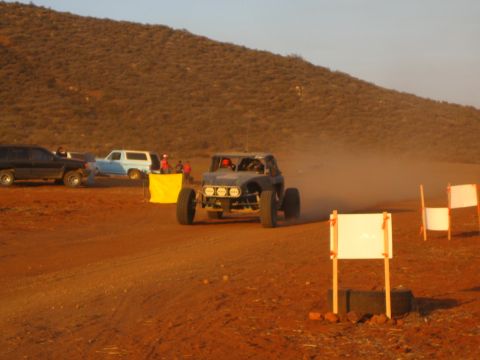 1990 Ford Bronco - off-road buggy fitted with a roof light bar approaches course markers on a desert dirt track as spectators and trucks wait nearby.