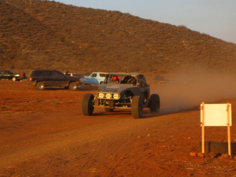 1990 Ford Bronco - off-road buggy with a row of round driving lights speeds along a dusty desert course past parked SUVs and a marker board.