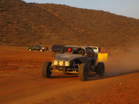 1990 Ford Bronco - two-seat off-road buggy with a front light bar kicks up dust on a desert track as spectators and a yellow marker line the route.