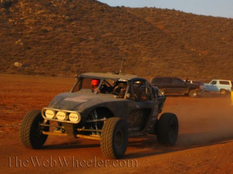 1990 Ford Bronco - off-road buggy fitted with round PIAA driving lights powers through a desert turn as parked SUVs sit along the hillside.