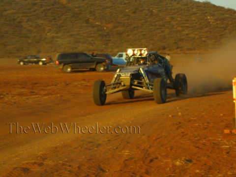 1990 Ford Bronco - open-frame off-road buggy with a roof-mounted light bar races along a dirt track as a line of trucks and SUVs sits parked behind.