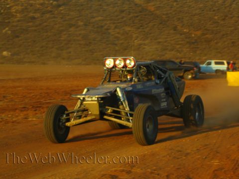 1990 Ford Bronco - blue open-tube off-road buggy with three round driving lights speeds along a desert dirt course toward a yellow course marker.