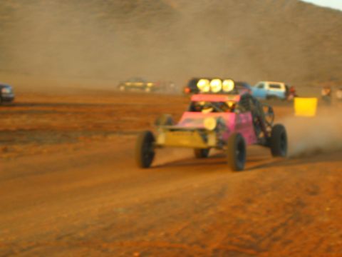 1990 Ford Bronco - pink and gold off-road buggy marked 999 races through dust on a desert course with other vehicles and a marker in the background.