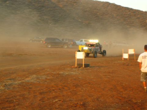 1990 Ford Bronco - off-road buggy with headlights blazing emerges from dust past white course markers as a spectator in white stands at right.