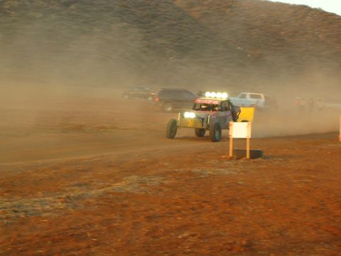1990 Ford Bronco - off-road buggy with bright headlights drives through thick dust on a desert track beside a marker board and parked vehicles.
