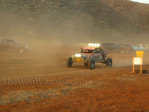 1990 Ford Bronco - off-road buggy with a roof light bar races through swirling dust on a desert course past parked trucks and a yellow marker.