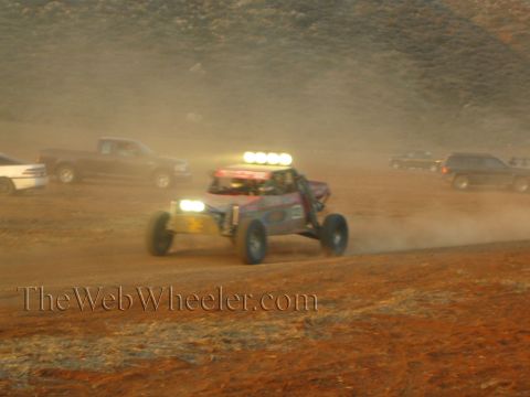 1990 Ford Bronco - off-road buggy with front and roof lights illuminated races through heavy dust on a desert course lined with parked vehicles.