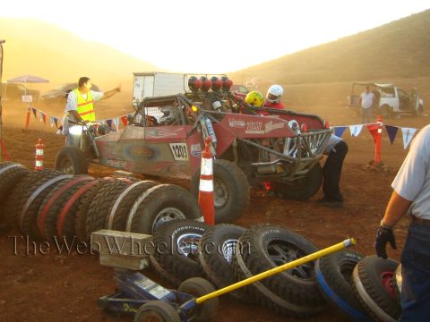 1990 Ford Bronco - red off-road race buggy sits in a dusty pit area surrounded by stacks of tires, crew members, and a flagged enclosure at golden hour.