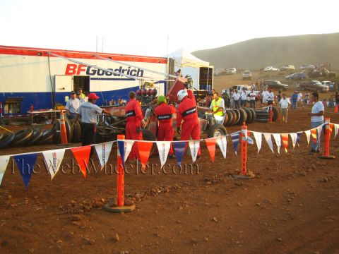1990 Ford Bronco - Crew in red suits service an off-road car beside a BFGoodrich support trailer in a desert pit ringed by red, white and blue pennants.