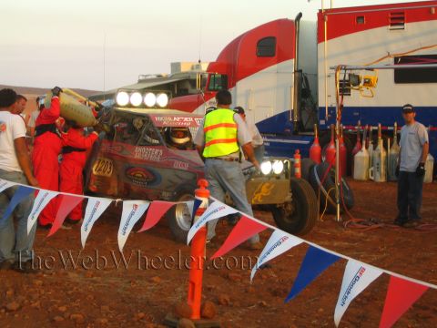 1990 Ford Bronco - pit crew works on a numbered off-road car with lights on, beside a red and blue support semi and BFGoodrich-branded pennants.
