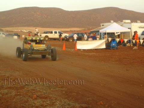 1990 Ford Bronco - yellow off-road buggy speeds along a desert dirt track near pop-up canopies, parked pickups, and trailers at golden hour.