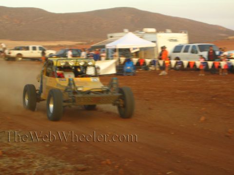 1990 Ford Bronco - yellow off-road buggy with driving lights corners on a desert track beside pop-up canopies, trailers, and spectators.