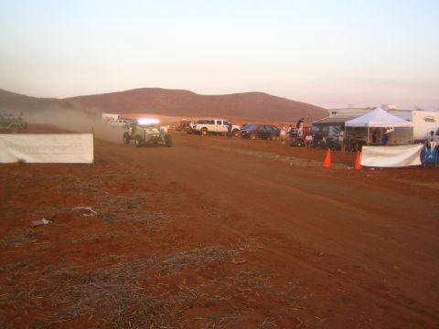 1990 Ford Bronco - off-road buggy with headlights on drives a hazy desert track at dusk past traffic cones, canopies, and a busy pit area.