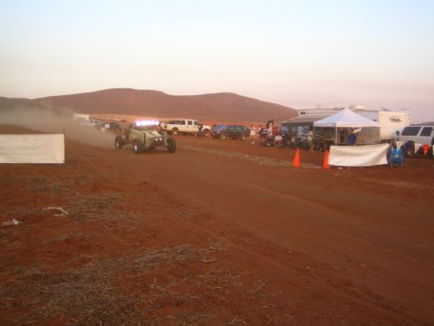 1990 Ford Bronco - off-road buggy with bright lights moves along a dusty desert track at twilight beside cones, a canopy, and parked vehicles.