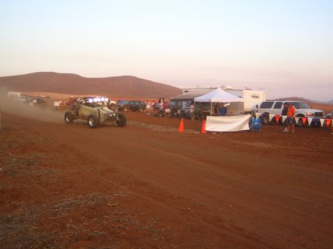 1990 Ford Bronco - off-road buggy with a lit light bar travels a desert track at dusk past traffic cones, a pit canopy, and parked SUVs and trailers.