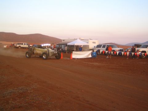 1990 Ford Bronco - single-seat off-road race buggy speeds along a red-dirt desert course at dusk past spectators, RVs and pickup trucks.