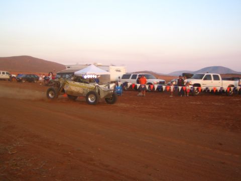 1990 Ford Bronco - tubular off-road race buggy races down a packed-dirt desert course at dusk with a line of pickups and a row of pennant flags alongside.