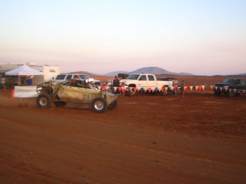 1990 Ford Bronco - Off-road race buggy number 1219 speeds along a red-dirt desert course past parked pickup trucks and pennant flags at dusk.