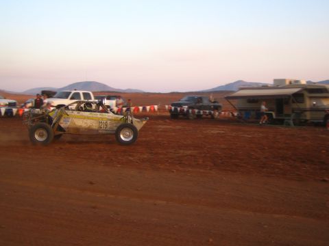 1990 Ford Bronco - Off-road race buggy number 1219 races across a flat red-dirt desert course past pickups and an RV at dusk.