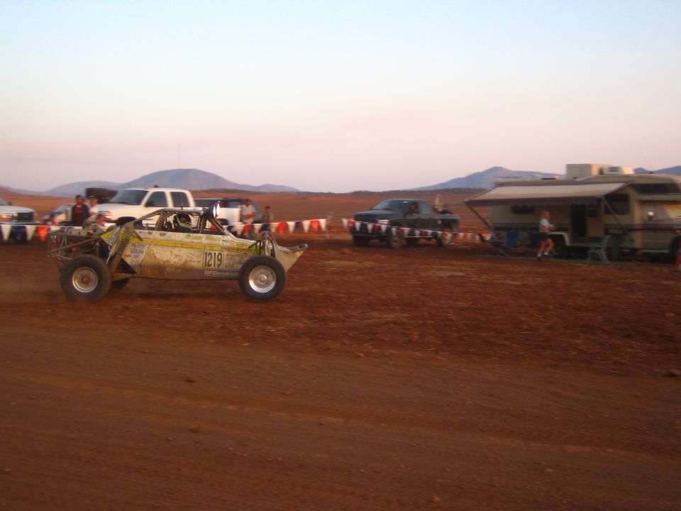 Off-road buggy 1219 racing across a desert course past an RV at dusk