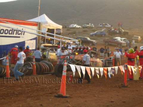 1990 Ford Bronco - Crew members in red suits work around off-road race vehicles in a dusty desert pit area with cones, pennant flags and a BFGoodrich tent.