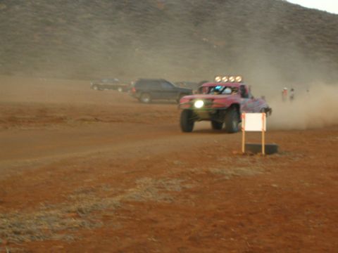 1990 Ford Bronco - off-road race truck with a roof light bar drives through swirling dust on a desert course past a parked SUV.