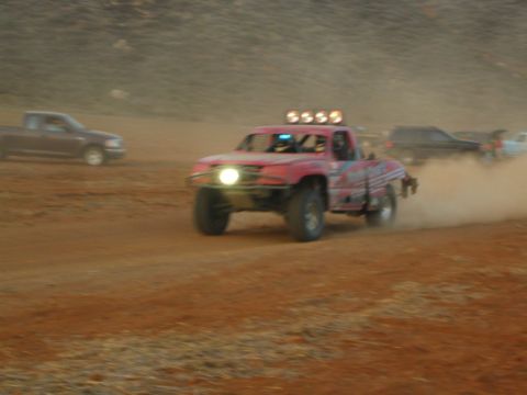 1990 Ford Bronco - pink off-road race truck with headlights and a roof light bar charges through dust on a desert course past parked vehicles.