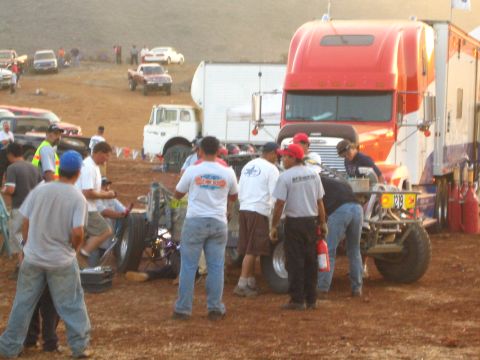 1990 Ford Bronco - pit crew in matching shirts works on an off-road race car in a dusty paddock beside a red semi-truck hauler.