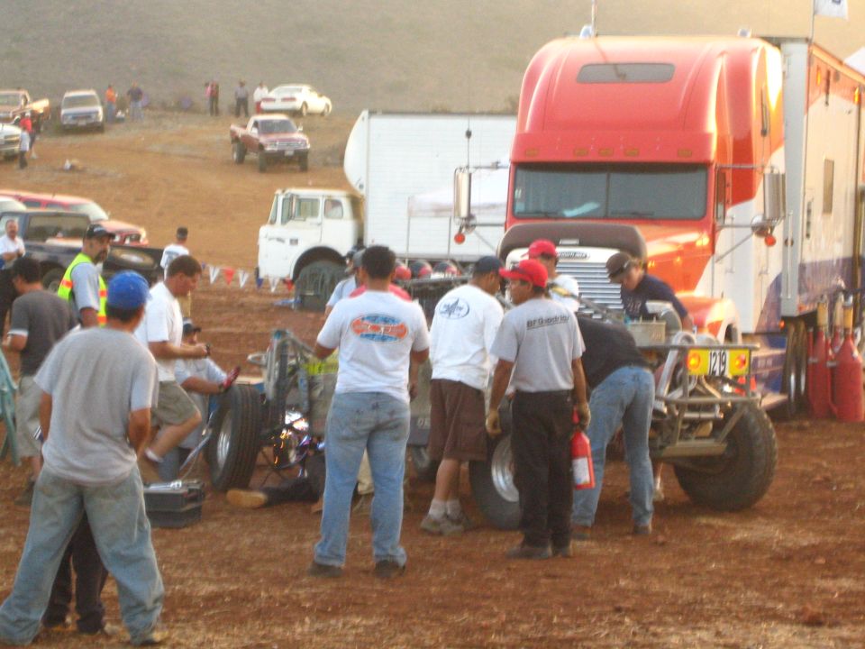 Pit crew working on an off-road race car beside a red semi-truck hauler