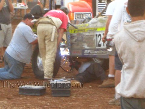 1990 Ford Bronco - Crew members weld on a yellow off-road race car numbered 121 with BFGoodrich livery as sparks fly in a dirt pit.