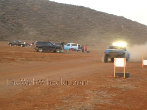 1990 Ford Bronco - blue off-road race truck with a glowing roof light bar kicks up dust on a desert course past parked trucks at dusk.