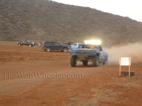 1990 Ford Bronco - blue off-road race truck with an illuminated light bar races across a red-dirt course past a black Jeep and pickups at dusk.
