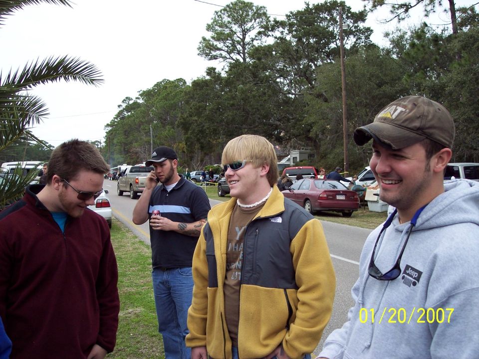 Group of men standing by a road with parked cars and trees in the background
