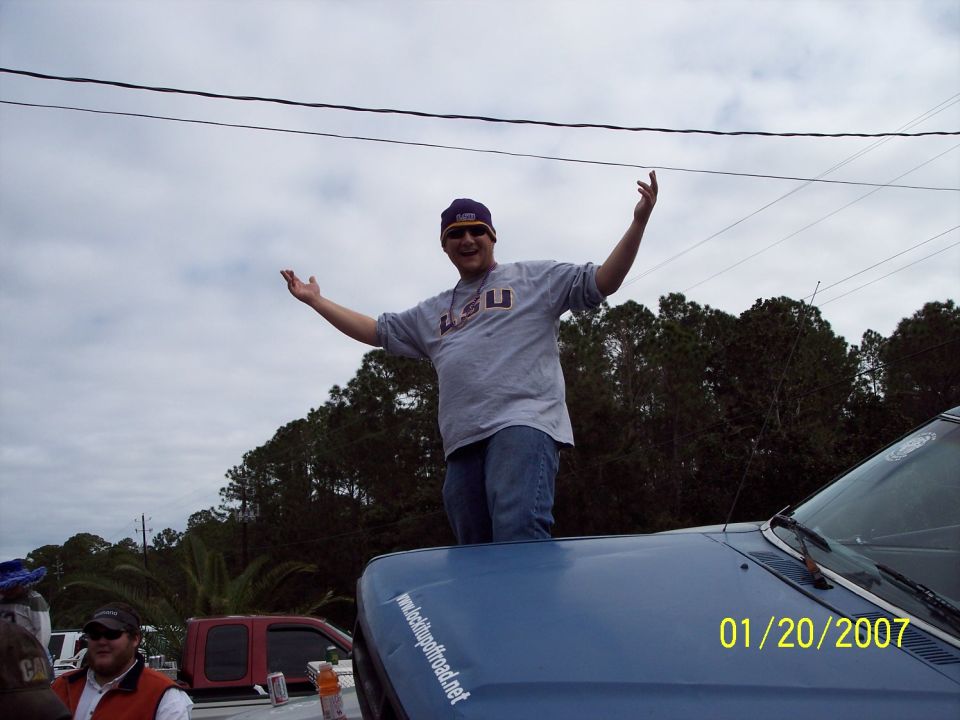 Man in LSU shirt stands on blue vehicle hood. Other vehicles and trees in background. Date stamp 01/20/2007