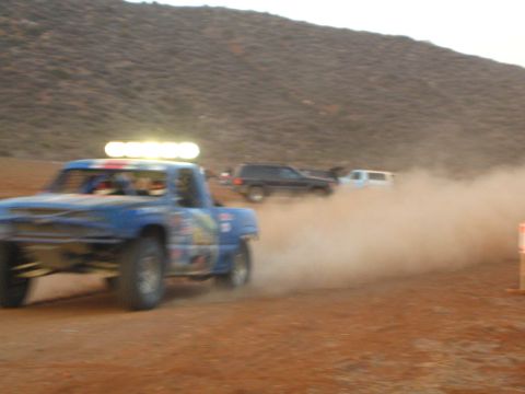 1990 Ford Bronco - blue off-road race truck with a roof light bar speeds through heavy dust on a desert course below a hillside.